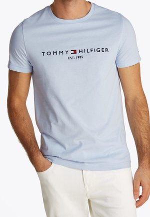 Mand iført lyseblå Tommy Hilfiger t-shirt med logo, flag og teksten "EST. 1985", kombineret med hvide bukser.