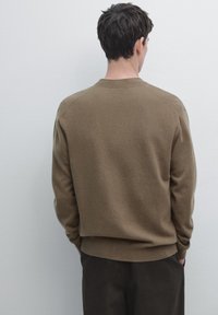 Brown, lightweight sweater with a crew neck, ribbed cuffs, and hem. Soft texture with a relaxed fit. Worn over dark trousers.