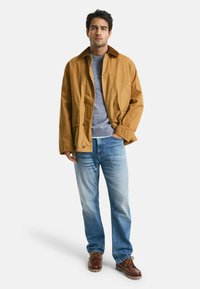 Beige Baumwolljacke mit großen Knöpfen, blauer Pullover darunter, hellblaue Jeans und braune Schnürschuhe. Steht vor einem weißen Hintergrund.