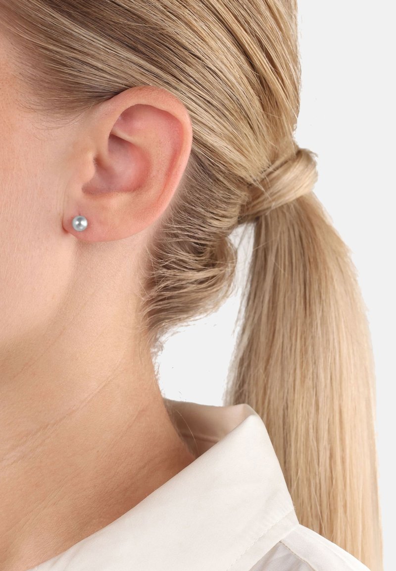 Donna bionda con capelli legati, che indossa piccoli orecchini a cerchio d'argento e una camicia bianca con colletto, su uno sfondo bianco.