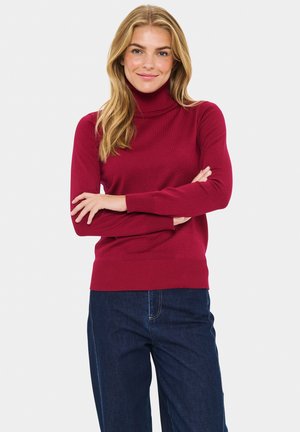 Pull à col roulé rouge en tissu tricoté doux, avec des manches longues et un ourlet côtelé. Porté avec un jean bleu foncé.