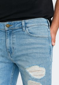 Pantalones vaqueros de mezclilla azul claro con un acabado desgastado, que presentan áreas desgastadas y un diseño clásico de cinco bolsillos. Incluyen un cierre de botón dorado.