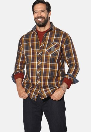 Braun-karierte Hemd mit Senfgelb und weißen Akzenten, mit Brusttasche und hochgekrempelten Ärmeln über einem roten Unterhemd. Dunkle Jeans sind sichtbar.