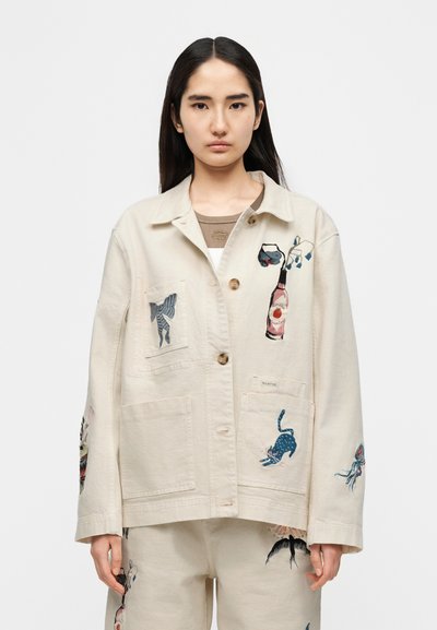 Μπεζ τζάκετ από καμβά με πολύχρωμα κεντημένα σχέδια ζώων και αντικειμένων, όπως ένα μπουκάλι και μια γάτα, με εμπρόσθιες τσέπες και κουμπιά.