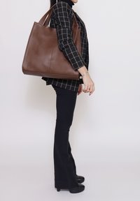 Borsa tote in pelle marrone con texture liscia, forma rettangolare larga e doppi manici, indossata da una persona con blazer nero a quadri.