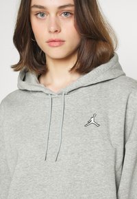 Grå hettegenser med snor, myk tekstur og en svart brodert Jumpman-logo på brystet. Enkel, uformell design.