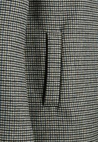 Närbild av en ficka med lock på en jacka i houndstooth-mönster i svarta, vita och beige nyanser.