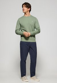 Ljusgrön sweatshirt med rund halsringning, ribbade ärmslut och nederkant. Mörk marinblå byxor, beige sneakers. Minimalistisk design med subtil branding.