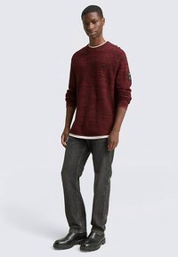 Vinröd texturerad tröja med långa ärmar och rund halsringning, lager-på-lager över en vit skjorta, ihop med mörka jeans och svarta ankelstövlar.