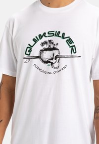 Camiseta blanca de algodón con un diseño gráfico que presenta una calavera, palmeras y una tabla de surf. El texto dice "Quiksilver Boardriding Company."