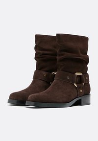 Bottines en cuir suédé marron avec un design froissé, présentant une large bande et des accents en métal doré. Talon carré et surface texturée.