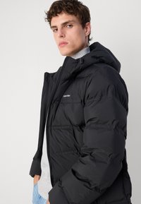 Zwarte pufferjack met ritssluiting, verstelbare capuchon en opvallende quiltpatronen. Logo op de linkerborst. G gedragen over een grijze trui.