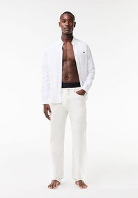 Chemise blanche à manches longues et boutons sur peau nue, associée à un pantalon couleur crème. Design ajusté, texture légère et détails minimalistes.