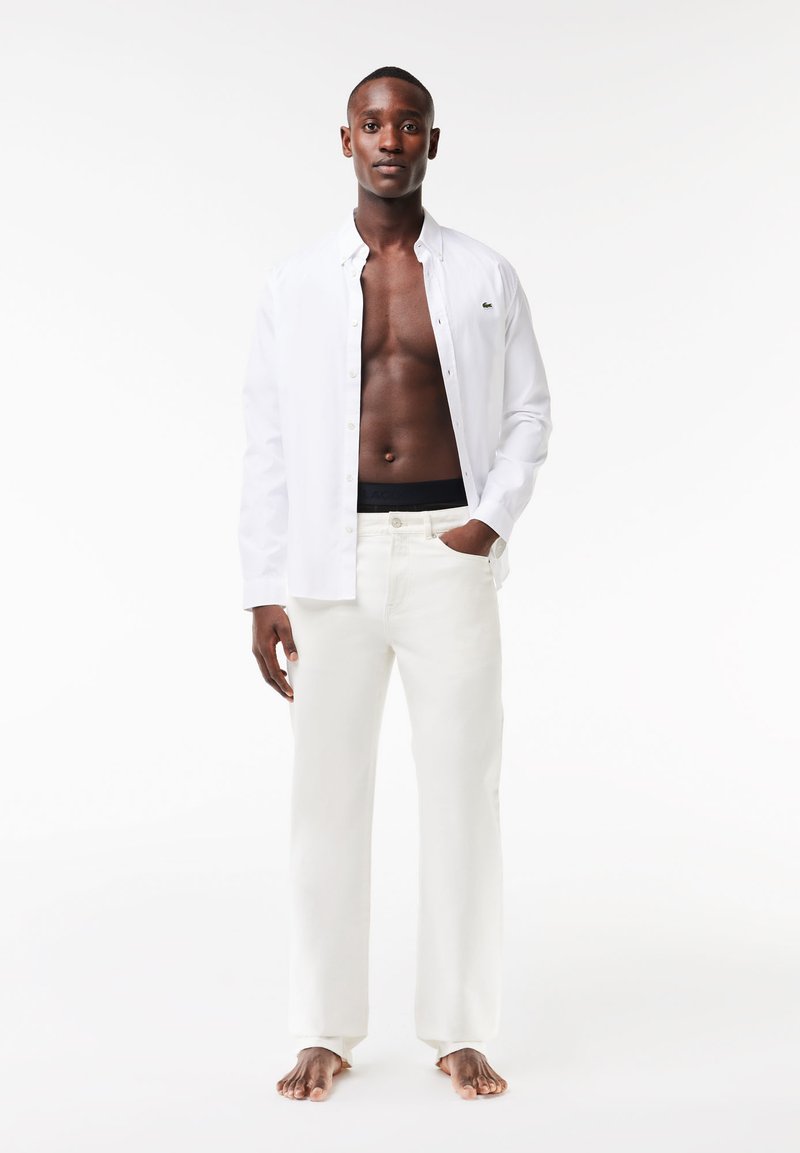 Weißes langärmliges Hemd mit Knöpfen über dem nackten Oberkörper, kombiniert mit cremefarbener Hose. Slim-Fit-Design, leichte Textur und minimalistische Details.