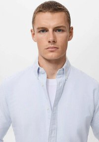 Camisa de botones a rayas azul claro y blanca con cuello abierto. Presenta puños abotonados y una tela texturizada. Se lleva sobre una camiseta blanca.