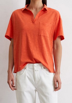 Person iført en løs orange kortærmet polo-shirt med lomme på brystet og hvide højtaljede jeans med dobbelte knapper.
