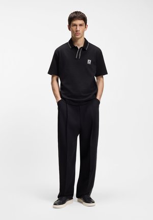 Male model wearing a black short-sleeve polo with logo and black wide-leg pants with side stripe, standing with hands in pockets against white background.