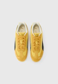 Un par de zapatillas Puma amarillas con rayas laterales y cordones negros, vistas desde arriba sobre un fondo blanco.