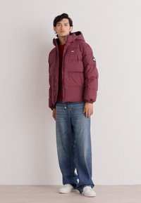 Maroon puffer jacket with a hood, featuring a zipper closure and logo patch on the sleeve. Worn over a red top and paired with loose blue jeans.