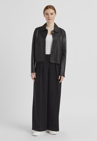 Schwarze Lederjacke in cropped Länge mit kleinem Kragen, zwei vorderen Taschen und Knopfverschluss, getragen über einem weißen gerippten Oberteil und lockeren schwarzen Hosen.