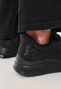 Melni sporta apavi ar teksturētu virsu un polsterētu apkaklīti. Iespējas ietver zemu profilu zoli un logotipa akcentu sānos.