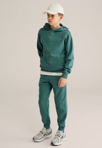 Tummanvihreä huppari, jossa on kengurutasku, yhdistettynä matching jogger-housuihin. Vaalean sävyinen lippalakki ja harmaat urheilukengät mustilla yksityiskohdilla täydentävät asun.