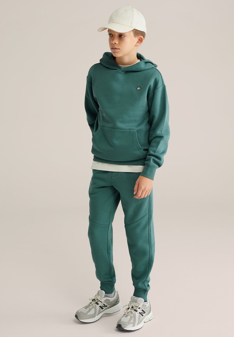 Tummanvihreä huppari, jossa on kengurutasku, yhdistettynä matching jogger-housuihin. Vaalean sävyinen lippalakki ja harmaat urheilukengät mustilla yksityiskohdilla täydentävät asun.