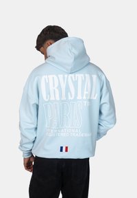 Felpa con cappuccio di colore azzurro, caratterizzata da un grande testo bianco "CRYSTAL PARIS" sul retro, con una piccola toppa della bandiera francese in basso.