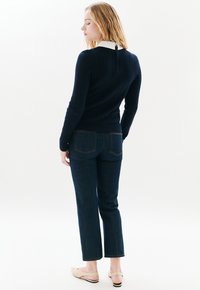Navyblauer Kabelstrickpullover mit einem weißen Kragen und einem Reißverschluss auf der Rückseite. Kombiniert mit dunkelblauen, geraden Jeans und beigen Schlupfschuhen.