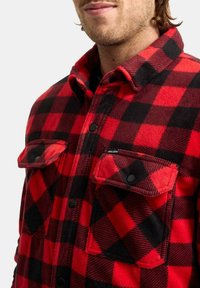 Camisa de franela a cuadros rojos y negros con cierre de botones, bolsillos en el pecho y cuello puntiagudo; textura suave y detalles de costuras visibles.