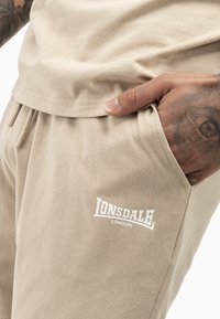 Pantaloni scurți bej din bumbac cu talie elastică și șnur, având un buzunar lateral și un logo alb "Lonsdale London" pe coapsa stângă.