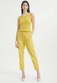 Żółta bezrękawnik i dopasowane spodnie, oba wykonane z gładkiego materiału. Modelka nosi beżowe buty na wysokim obcasie. Prosty, elegancki design.
