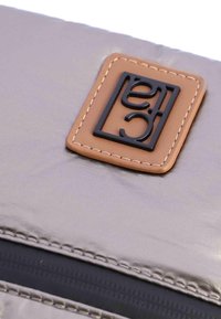 Bolso plateado metálico con logotipo negro en una etiqueta de cuero marrón. Presenta detalles de costura y un cierre de cremallera. Textura suave.