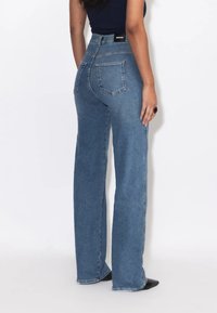 Frau trägt hochgeschnittene, gerade blaue Jeans und ein dunkles Oberteil, steht mit dem Rücken zur Kamera vor einem schlichten Hintergrund.