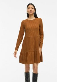 Robe en tricot côtelé marron avec des manches longues, un col rond et un ourlet à volants en couches. Le tissu a un aspect texturé et un léger éclat.