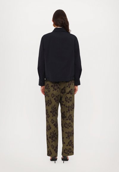 Zwarte jas met een rechte snit, gecombineerd met olijfgroene wijde broek met een donker bloemenpatroon. Dichte hakken.