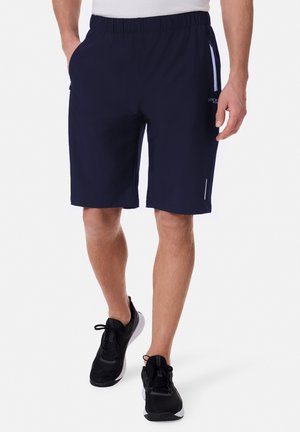 Mann trägt marineblaue Sportshorts mit Reißverschlusstaschen und schwarze Laufschuhe vor weißem Hintergrund.