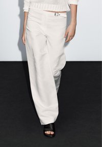 Witte wijde broek met een gladde structuur, met een sluiting met knopen aan de voorkant en riemlussen. Gecombineerd met zwarte sandalen met open tenen.