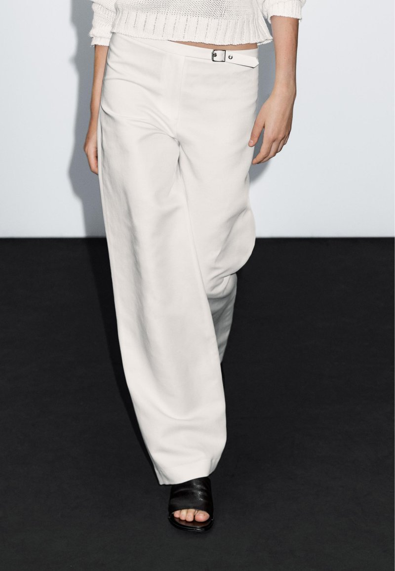Witte wijde broek met een gladde structuur, met een sluiting met knopen aan de voorkant en riemlussen. Gecombineerd met zwarte sandalen met open tenen.