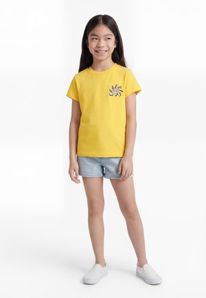 Dziewczyna z długimi ciemnymi włosami, ubrana w żółtą koszulkę z nadrukiem, jasnoniebieskie szorty jeansowe i białe buty wsuwane, stojąca i uśmiechająca się.