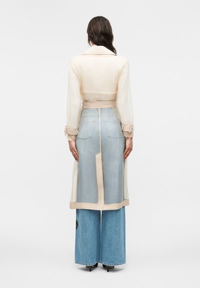Femme vue de dos portant un trench-coat beige transparent superposé sur un jean large bleu clair et des talons noirs sur un fond blanc.