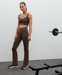 Brązowy zestaw sportowy składający się z dopasowanego topu odsłaniającego brzuch i spodni z wysokim stanem w kształcie dzwonów, wykonany z gładkiego materiału. Czarne buty sportowe. W pobliżu ławka do podnoszenia ciężarów.