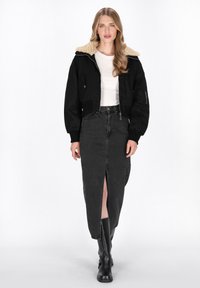 Schwarze, kurz geschnittene Jacke mit einem hellbraunen Schafslederkragen, kombiniert mit einem schwarzen Denim-Rock mit einem Schlitz vorne. Das Modell trägt schwarze kniehohe Stiefel.