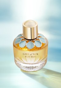 Rund glassparfymeflaske med en gull- og lys blå blomsterdetalj, med etikettene "GIRL of NOW" og "ELIE SAAB", fylt med ravfarget væske.