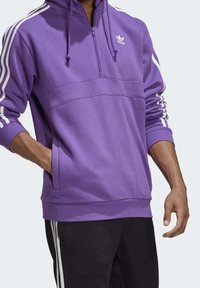 Lila zip-up hoodie med vita ränder längs ärmarna och en ficka framtill. Tillverkad av mjukt, texturerat material. Adidas-logotyp på bröstet.