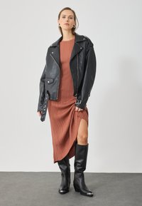 Černá kožená motorkářská bunda přes proplétanou midi šaty v zemských tónech s bočním rozparkem, doplněná koženými botami do výšky kolen.