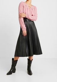 Blusa a maniche lunghe rosa chiaro con dettaglio a fessura, abbinata a una gonna midi plissettata nera. Stivali cowboy in pelle neri con tacco completano il look.