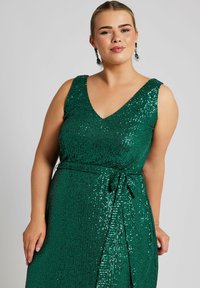 Grön paljettklänning med V-halsringning, ärmlös design, rynkad bodice och knytmidja. Har en glittrande, texturerad yta.