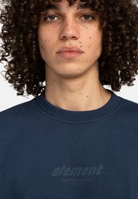 Navy blue sweatshirt with ribbed neckline features subtle embossed logo. Soft texture and relaxed fit. Model with curly hair is centered.