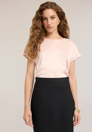 Mujer con el pelo largo y ondulado que lleva una camiseta de manga corta de color rosa claro, falda negra, pendientes de oro y una pulsera de oro, de pie frente a un fondo liso.